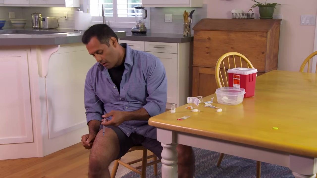 A person holding a syringe steady in their thigh at a kitchen table, with medical supplies and a sharps container nearby.