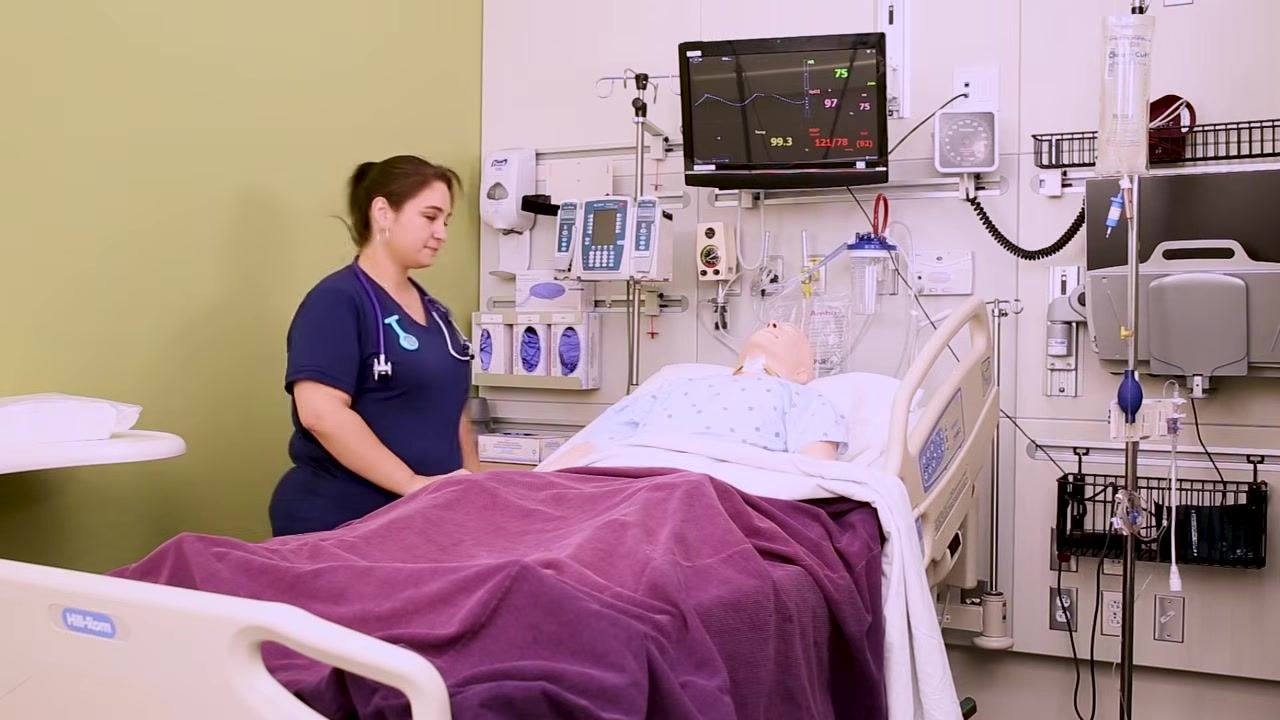 A nurse stands beside a hospital bed with a mannequin patient lying supine and covered with a blanket, in a clinical setting with medical equipment and monitors visible.