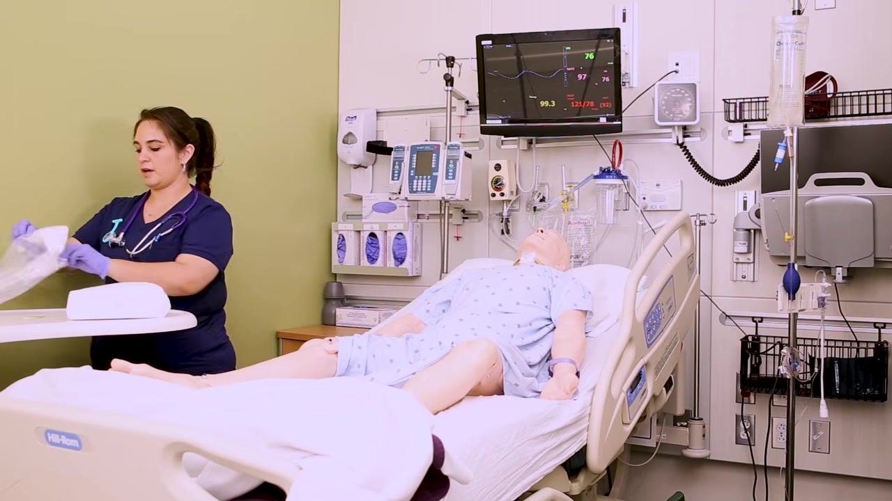 A nurse opens a plastic bag on a bedside table to prepare the Foley catheter kit, with the patient mannequin positioned with knees bent and legs apart.
