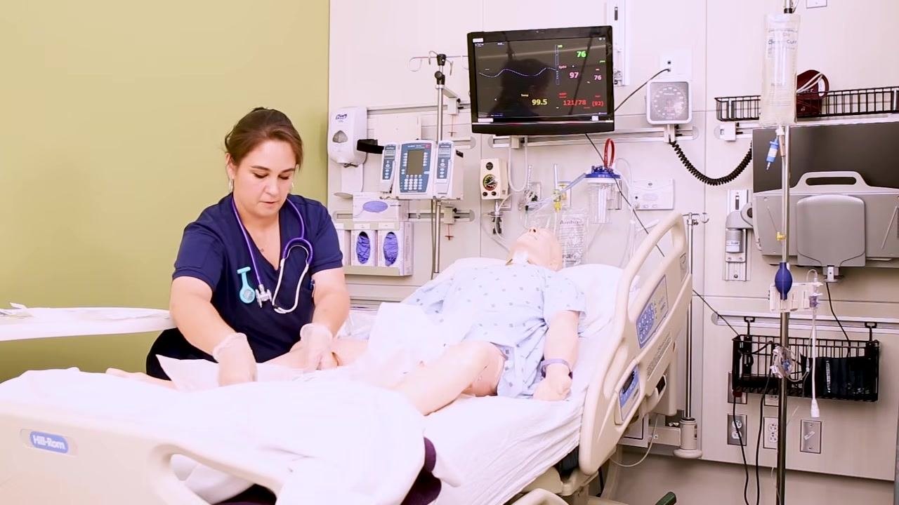 A nurse wearing sterile gloves carefully places a large sterile drape under the mannequin patient's perineum, ensuring her hands only touch the drape.
