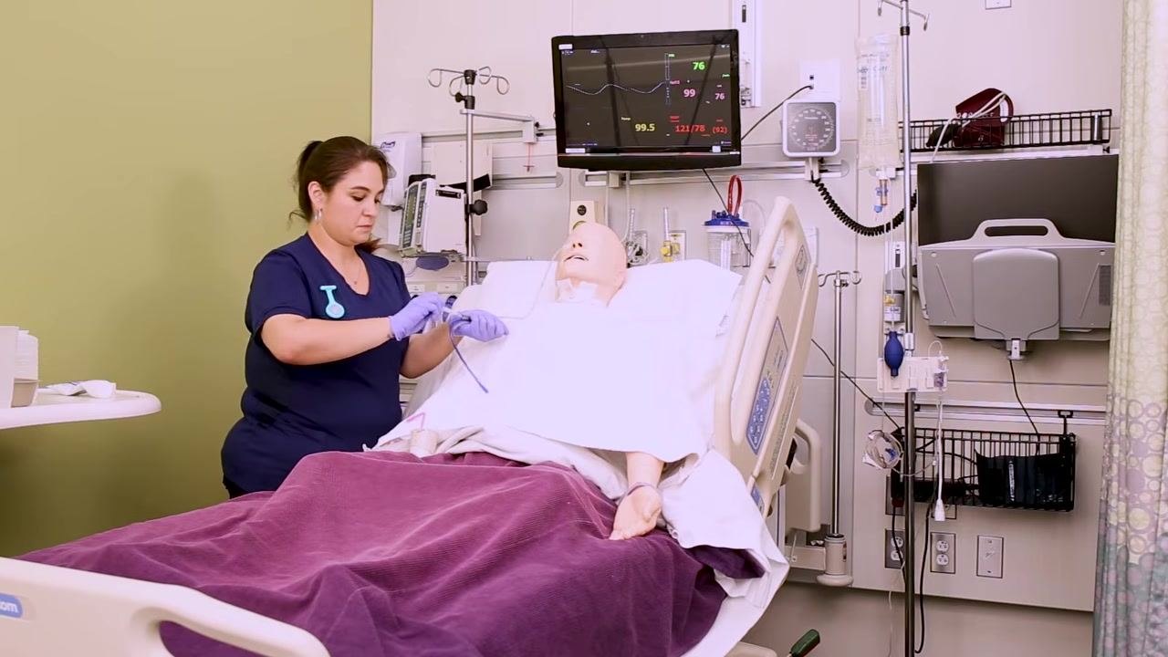 Nurse clamping and securing the NG tube to the mannequin's gown, finalizing the procedure, with medical monitors and equipment visible in the background