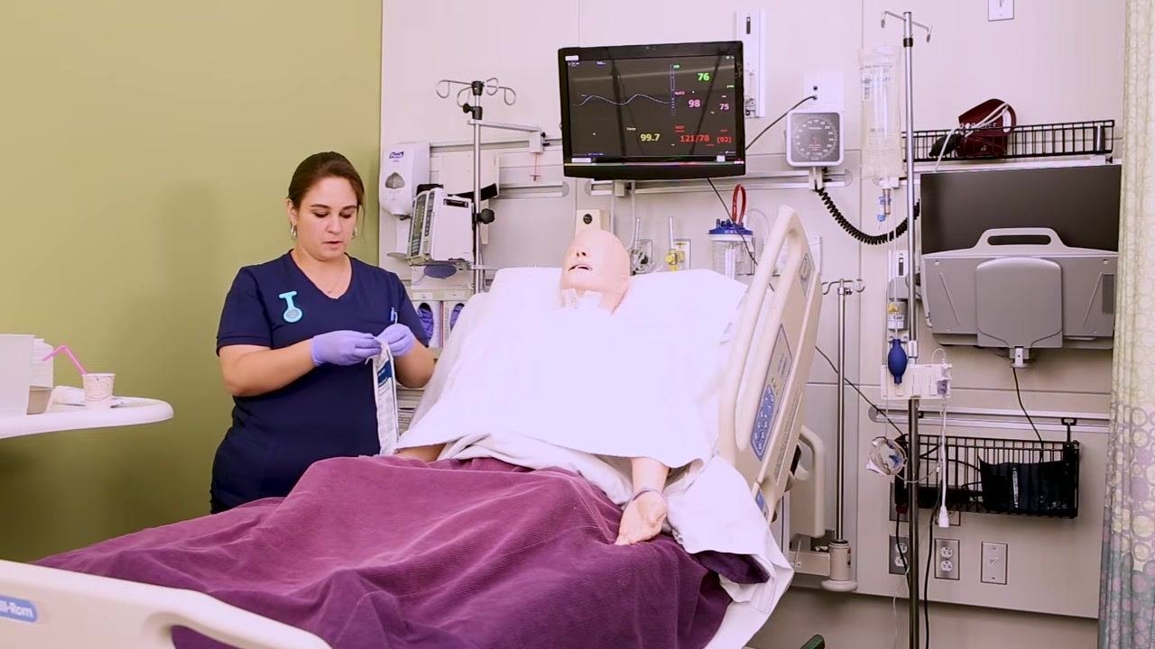 Nurse placing a towel across the chest of a patient mannequin in a hospital bed, with medical equipment and monitors in the background