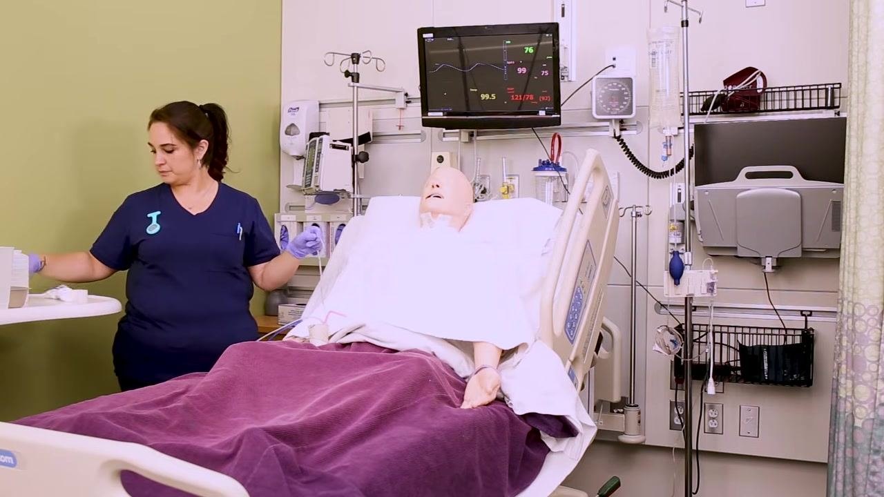 Nurse standing beside a patient mannequin in a hospital bed, preparing for NG tube insertion, with the mannequin upright, a towel on the chest, and medical monitors visible in the background