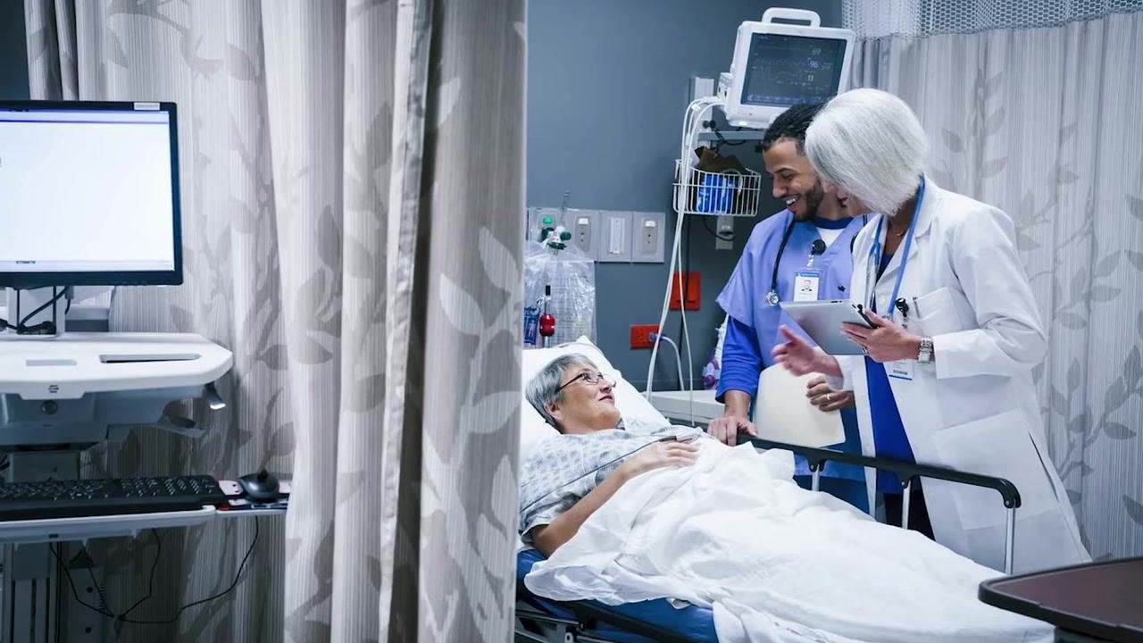 A healthcare worker wearing a mask and gloves holds a Foley catheter, showing its length and structure.