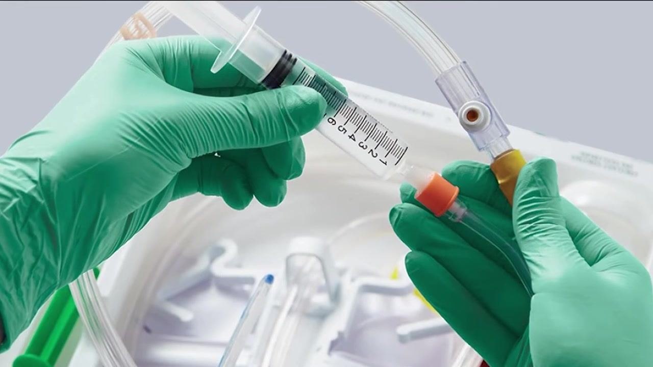 A person wearing green gloves holds a syringe and connects it to a Foley catheter tube, preparing to flush the catheter, with medical supplies visible in the background.