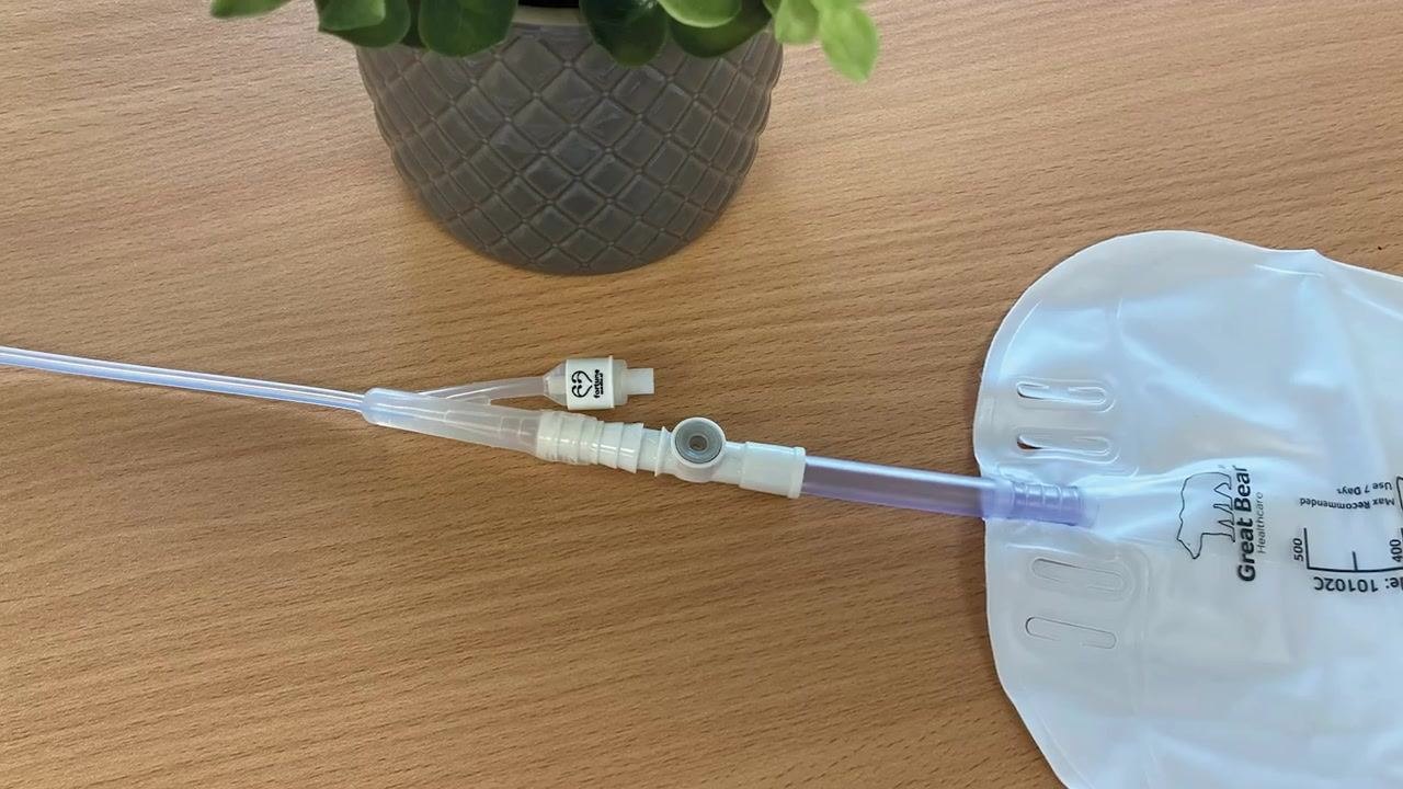 A Foley catheter drainage bag with the tubing disconnected and a white plug inserted into the open end, placed on a wooden surface next to a plant.