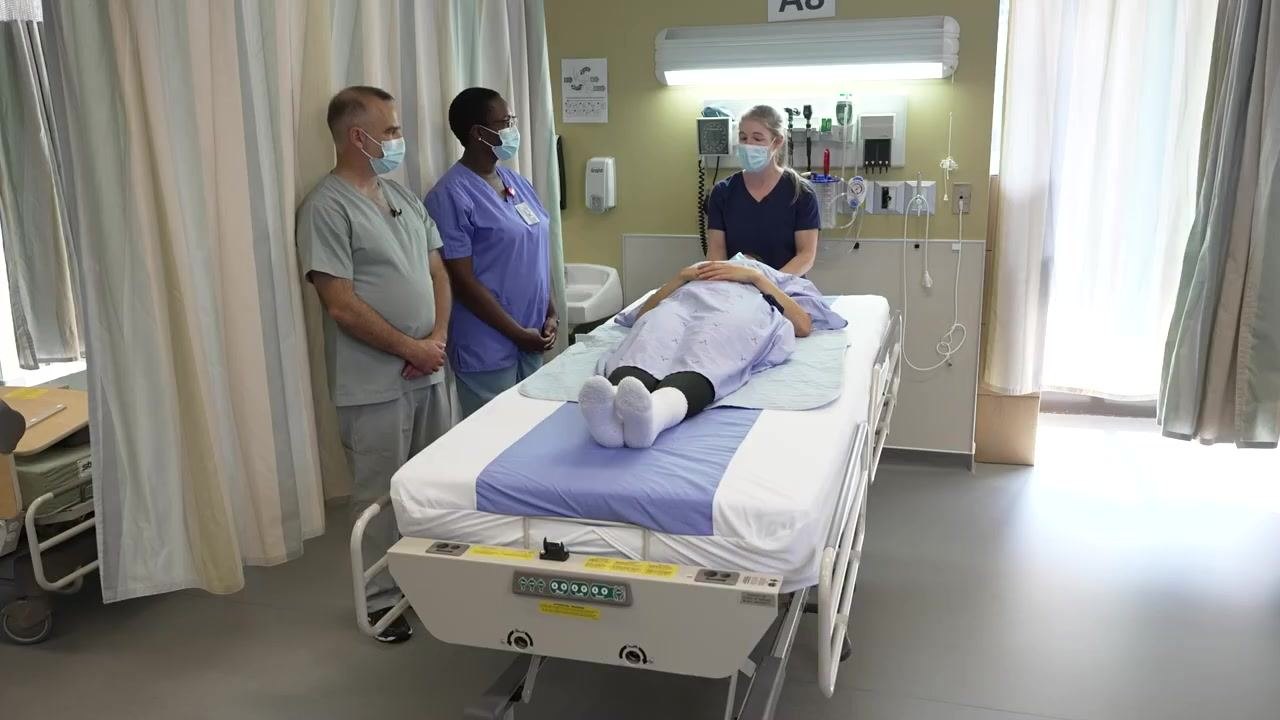 Two healthcare workers on the same side of the bed, gripping the patient at shoulder, hip, waist, and thigh while demonstrating proper body mechanics