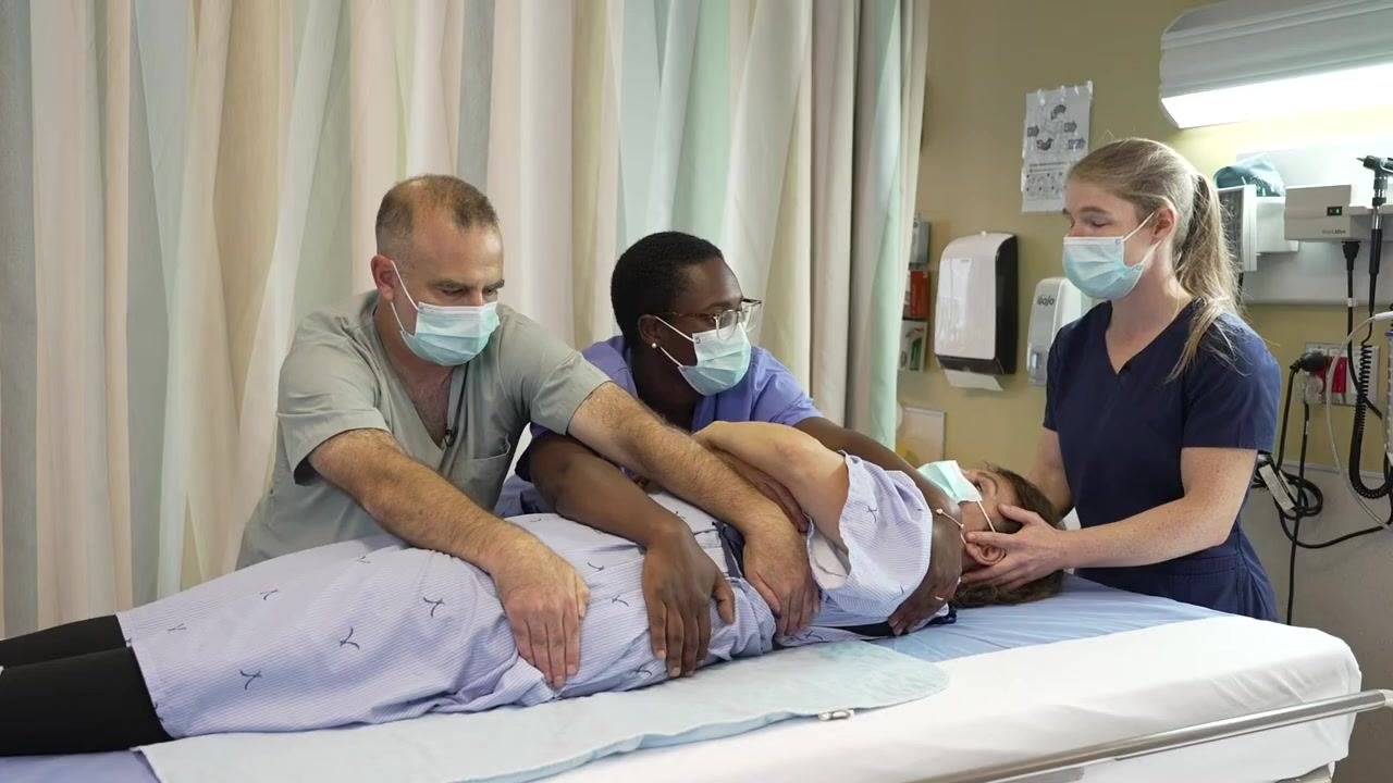 Three healthcare workers performing a forward-backward weight transfer, with one worker stabilizing the head and two supporting the body at the shoulder, hip, waist, and thigh