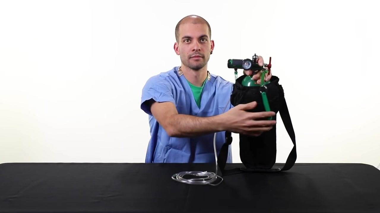 A person in scrubs holds an oxygen tank in a black carrier, adjusting the regulator and preparing to empty the tank, with nasal cannula tubing on the table.