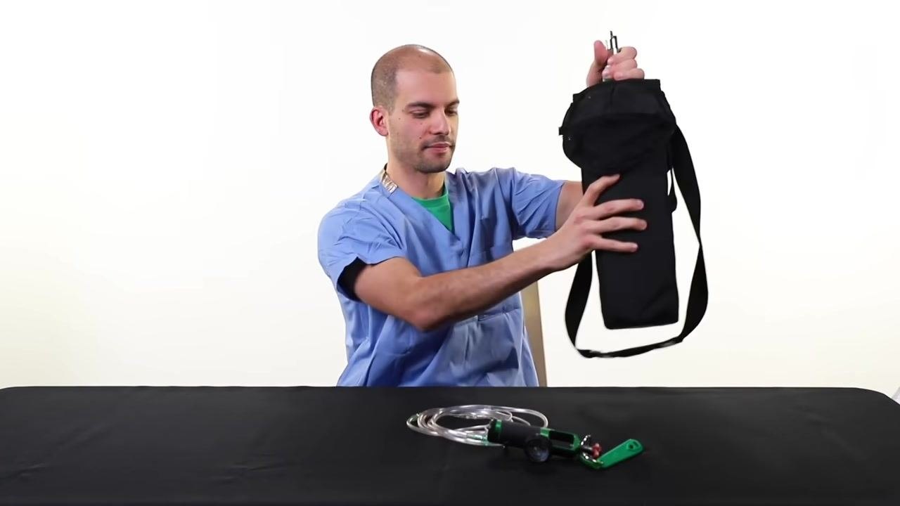 A person in scrubs lifts the empty oxygen tank in its black carrier off the table, with the regulator and tubing placed in front.