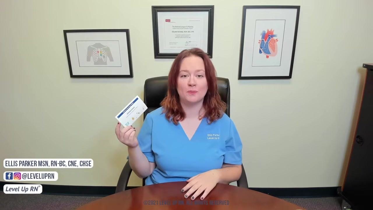 An instructor in blue scrubs holds the Clinical Nursing Skills Deck card while seated at a desk with framed medical illustrations and a certificate on the wall in the background.