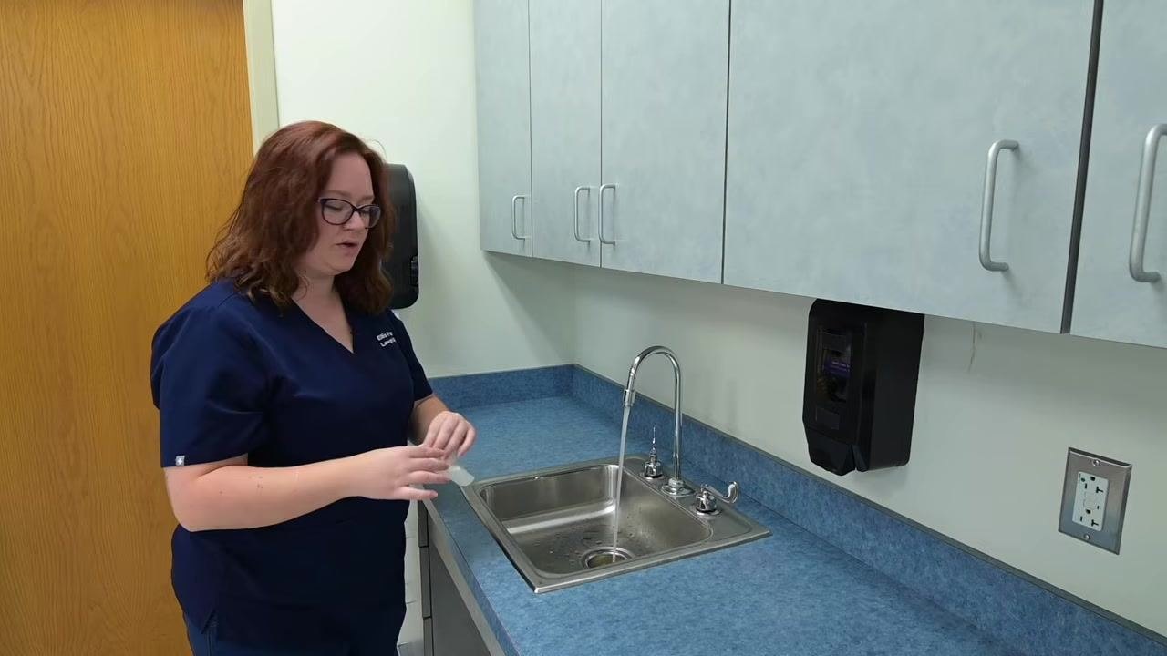 A nurse uses a paper towel to turn off the faucet at the clinical sink, avoiding direct contact with the handles.