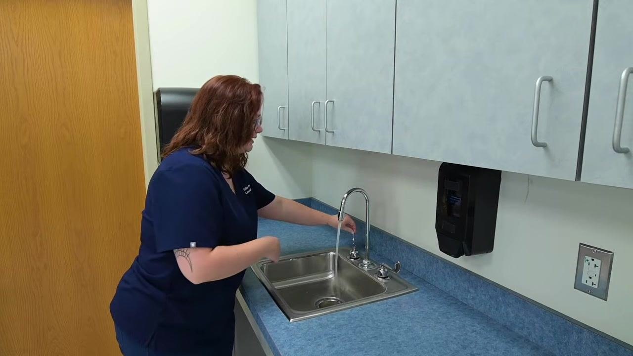 A nurse turns on the faucet at a clinical sink, positioning hands under the running water stream in preparation for washing.
