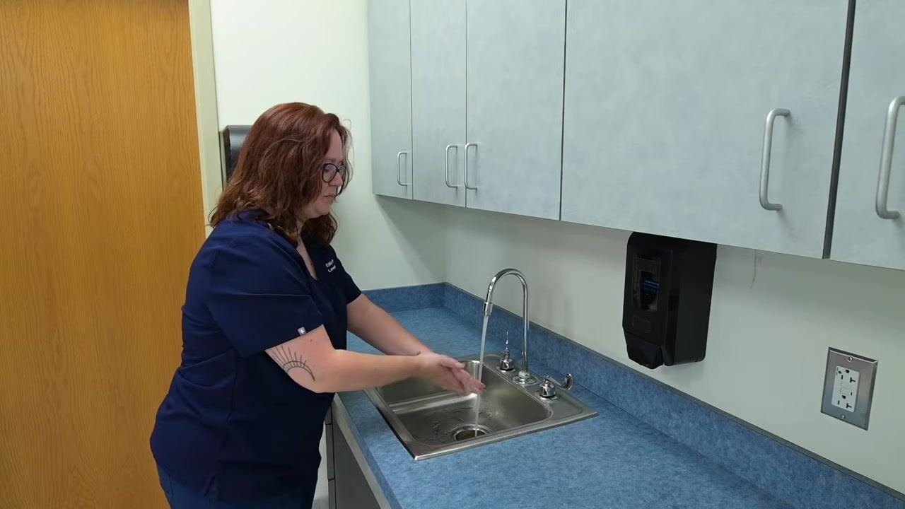 A nurse scrubs both hands together under running water at a clinical sink, demonstrating a thorough cleaning technique.
