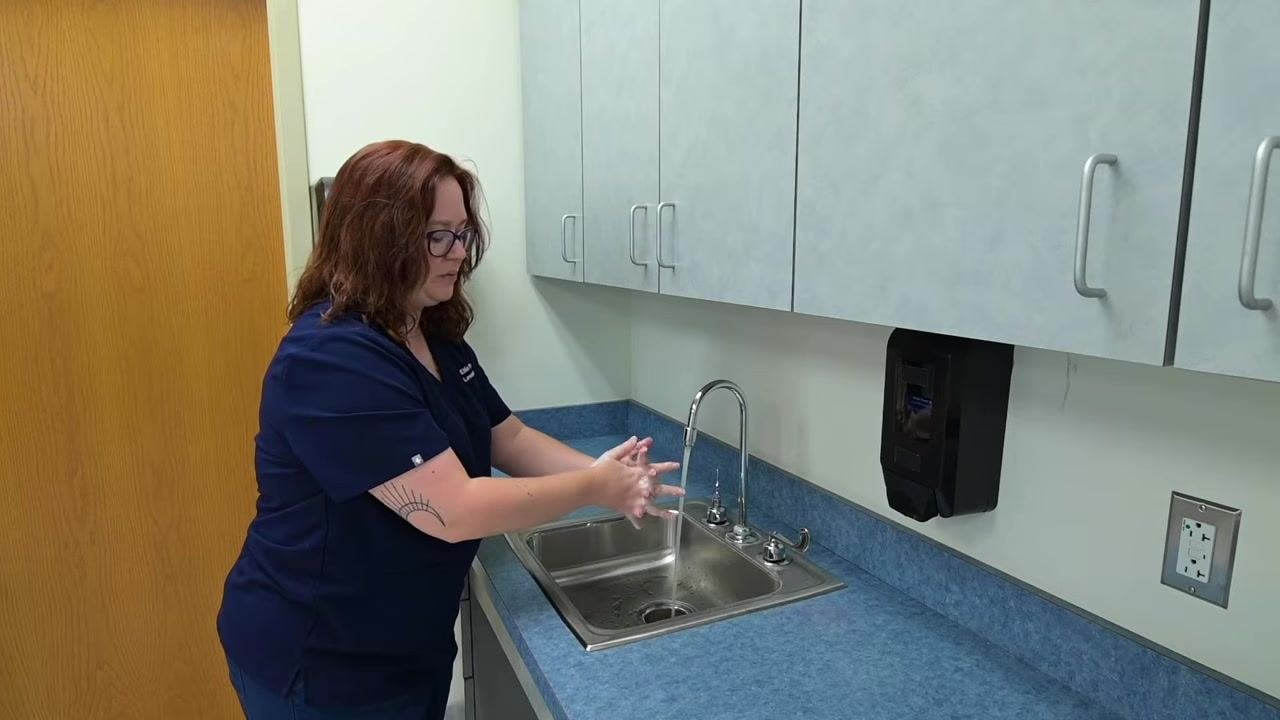 A nurse continues scrubbing hands, focusing on all surfaces while maintaining the correct downward hand position over the sink.