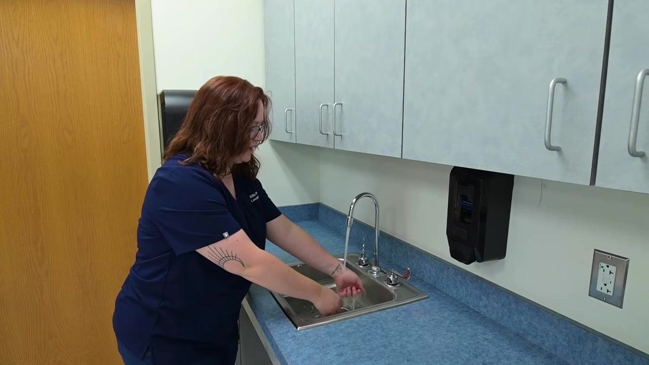 A nurse in navy scrubs rinses hands under running water at a clinical sink, keeping hands pointed downward and avoiding contact with the sink basin, with cabinets, a paper towel dispenser, and a soap dispenser visible nearby.