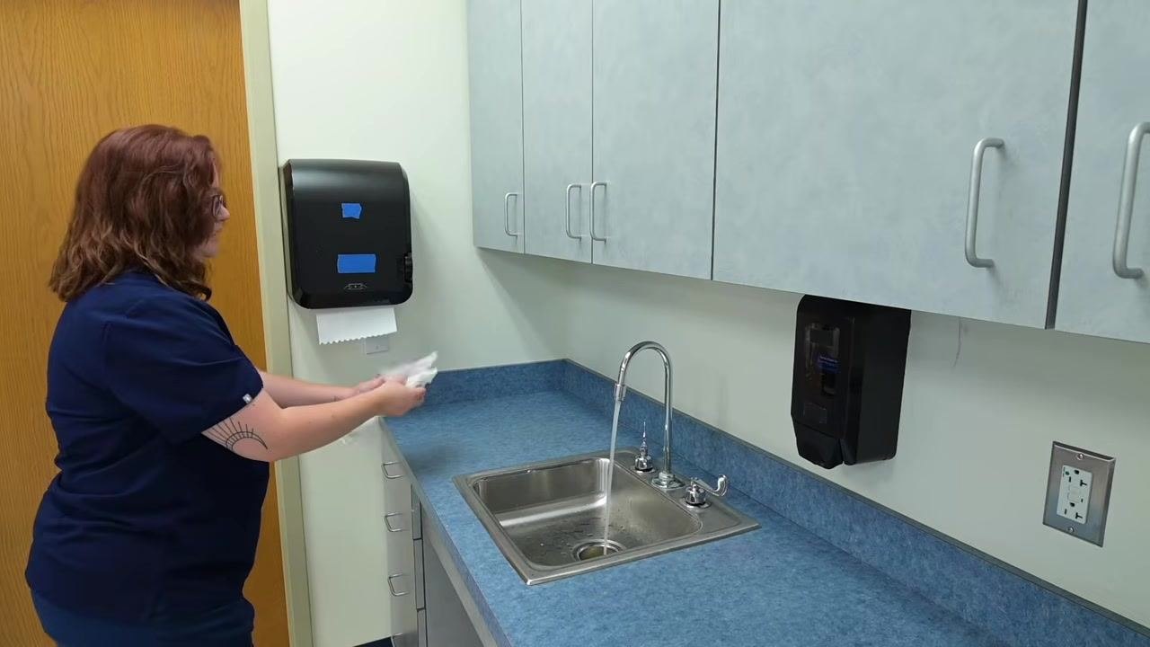 A nurse retrieves a paper towel from the dispenser in preparation for drying hands at the clinical sink, with the sink, faucet, and dispensers visible.