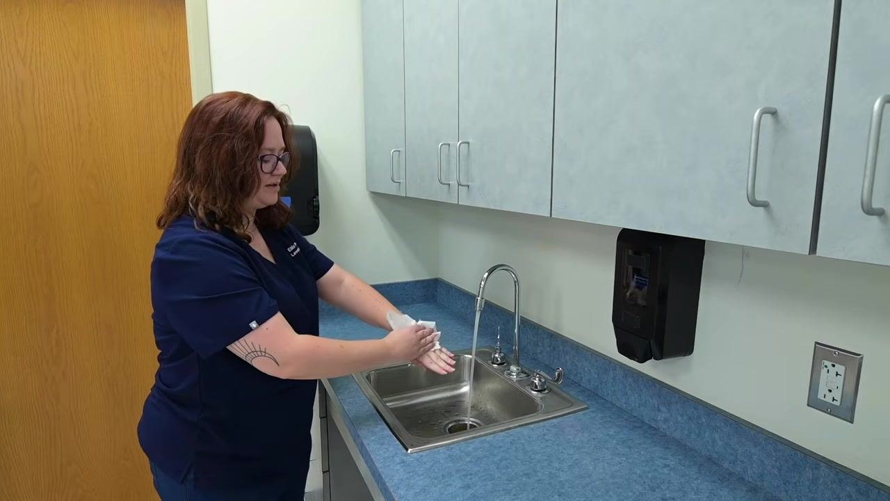 A nurse pats hands dry with a paper towel over the sink, demonstrating the correct drying technique.
