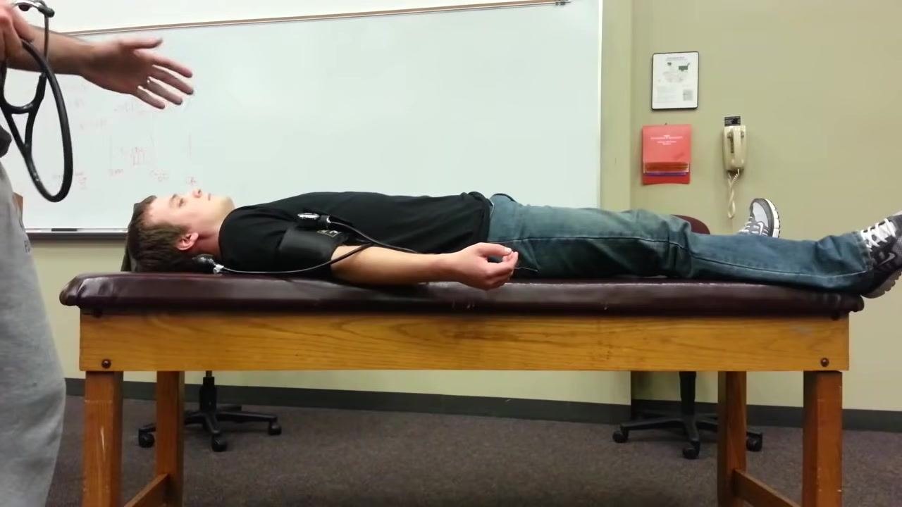Healthcare provider measuring the blood pressure of a patient lying on the table using a stethoscope and sphygmomanometer, with the patient relaxed and the cuff in place on the extended arm.