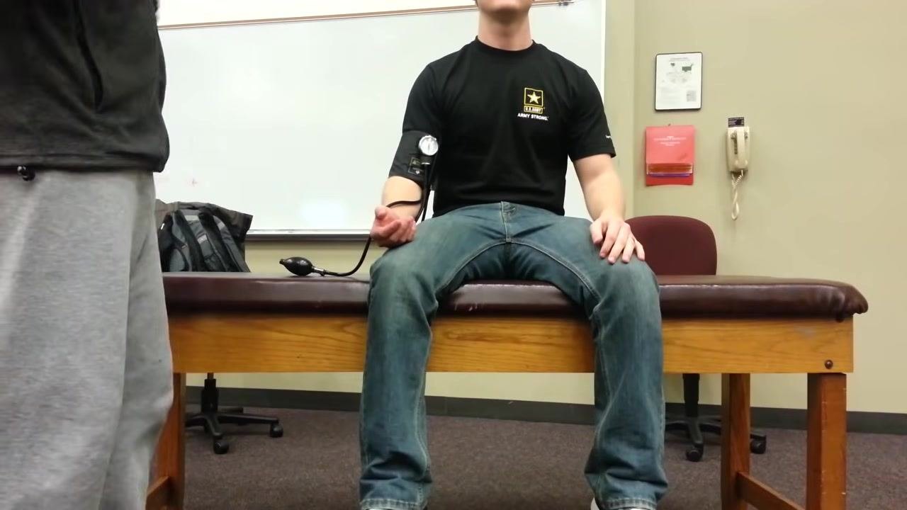 Patient sitting upright on the examination table with a blood pressure cuff on the left arm, holding the bulb, while a healthcare provider stands nearby preparing to take the next reading.