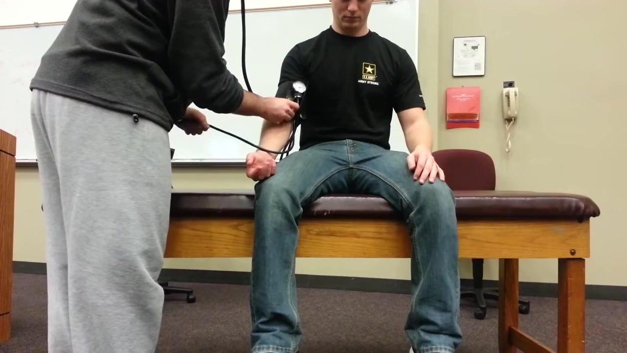 Patient sitting upright on the examination table with a blood pressure cuff on the left arm, while a healthcare provider holds the sphygmomanometer and stethoscope to take the reading.
