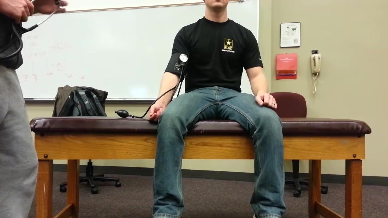 Patient standing upright next to the examination table with the blood pressure cuff still on the left arm, while a healthcare provider prepares to take the standing blood pressure reading.