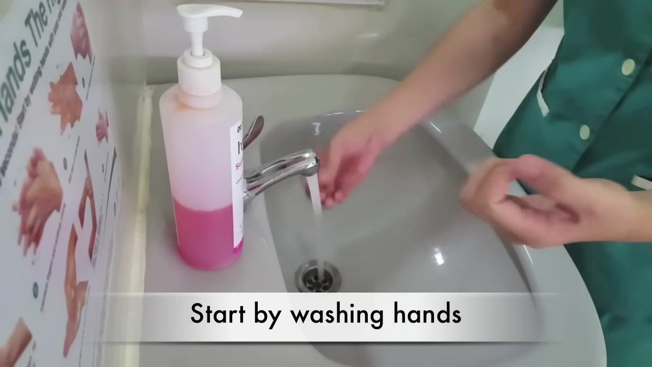 A person in a green uniform wets their hands under running water at a sink. A large bottle of pink liquid soap is on the left, and a handwashing instruction poster is visible on the wall. The caption reads "Start by washing hands."