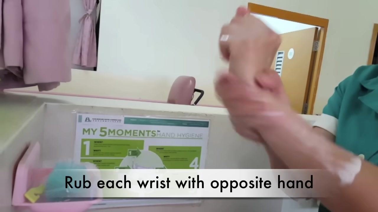A person rubs each wrist with the opposite hand, as indicated by the caption "Rub each wrist with opposite hand." A hand hygiene poster and cleaning supplies are visible in the background.