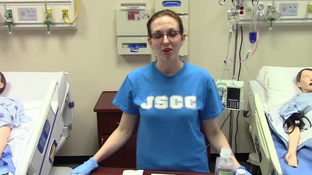 Clinician standing between two hospital beds, wearing gloves, with a trach care kit and supplies laid out on the table. Hospital environment with medical mannequins in beds.