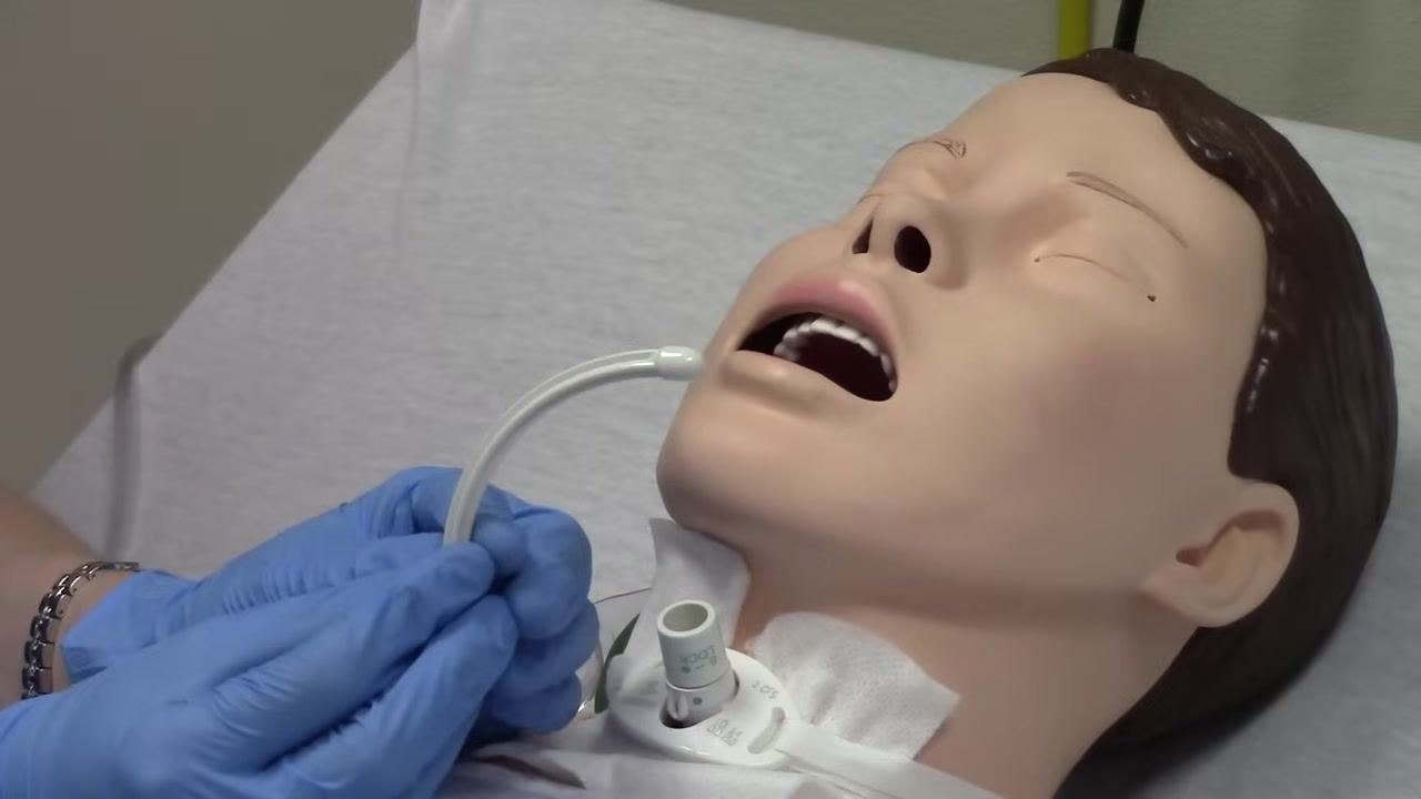 Gloved hands holding a white obturator, demonstrating its curved shape and use for reinserting the trach tube. The mannequin's trach site is visible.