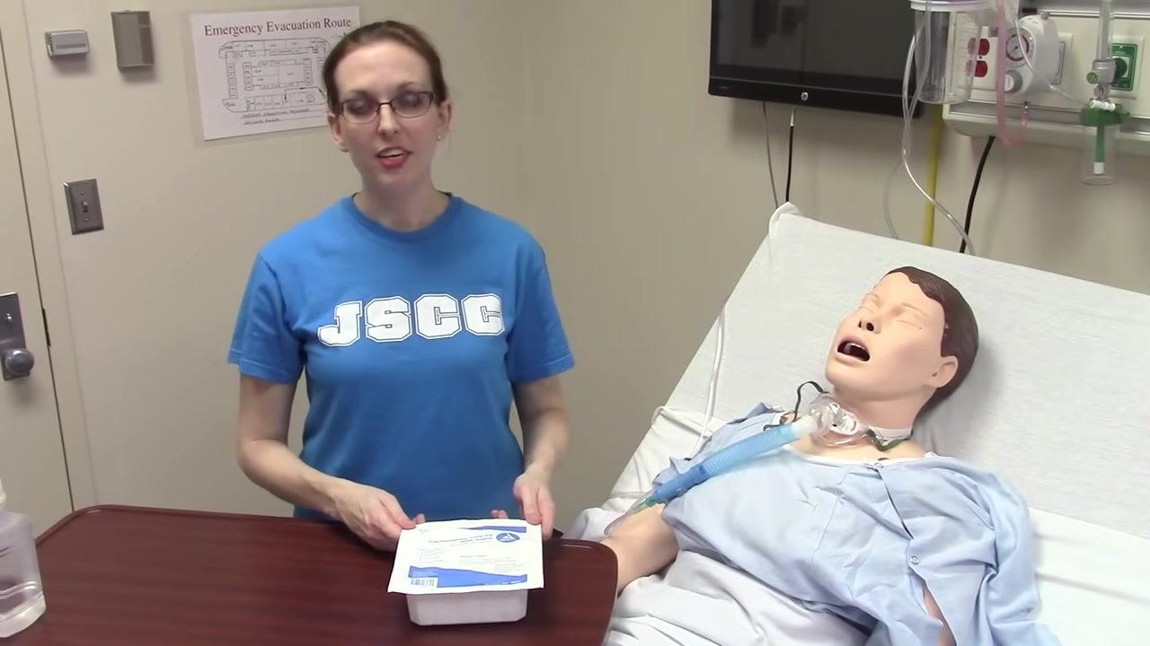 A clinician stands beside a hospital bed with a medical mannequin, holding a sealed trach care kit on a table and preparing to open it. The environment is a clinical simulation room with medical equipment and an emergency evacuation map on the wall.