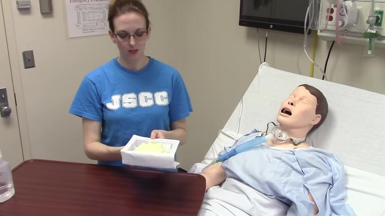 A clinician stands next to a hospital bed with a mannequin patient, holding a tray containing yellow sterile gloves and other supplies. The clinical room shows medical equipment and an emergency evacuation plan on the wall.
