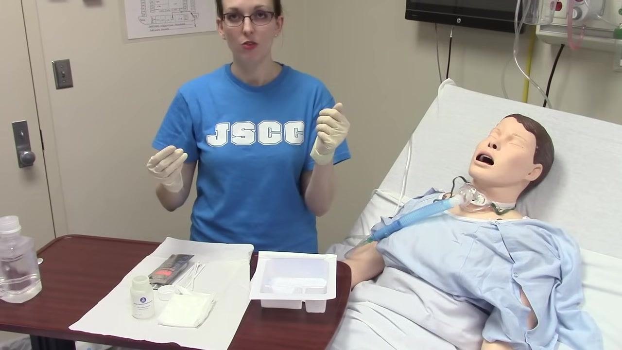 A clinician stands next to a mannequin patient on a hospital bed, wearing sterile gloves and holding both hands above a tray with medical supplies, preparing for the next step.