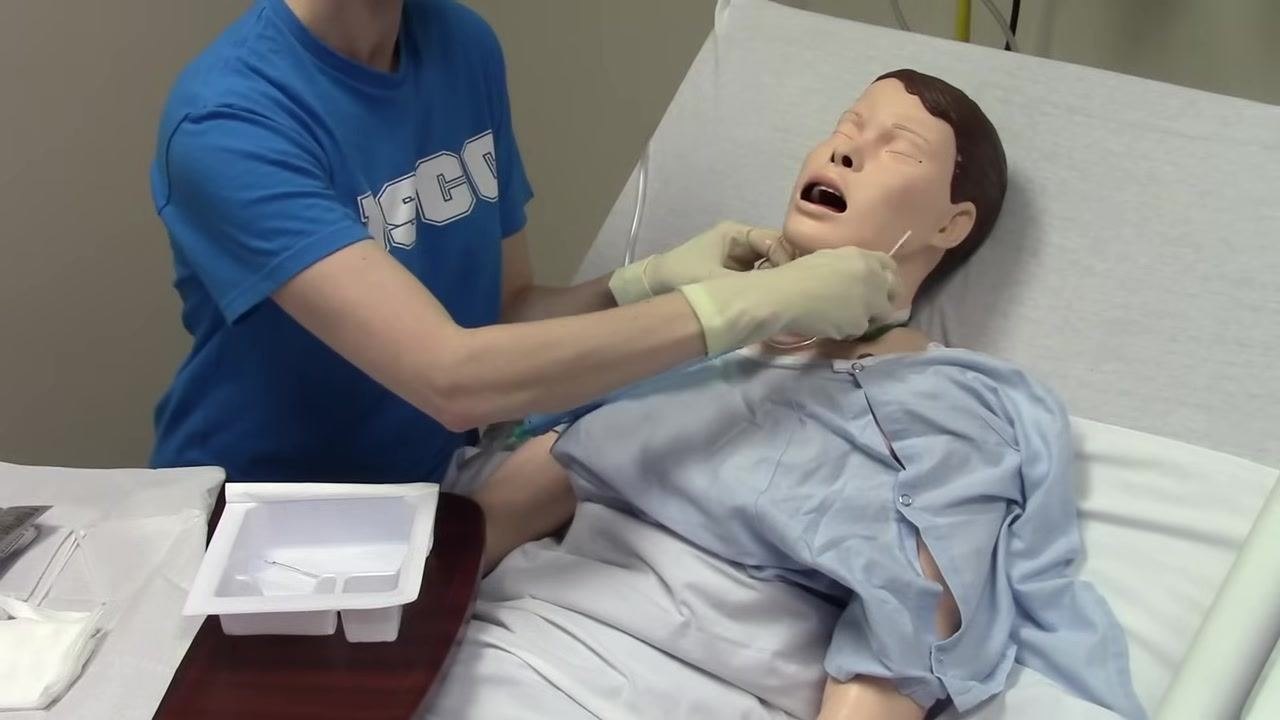 Gloved hands hold the inner cannula above the tray, preparing to return the oxygen mask to the mannequin patient.