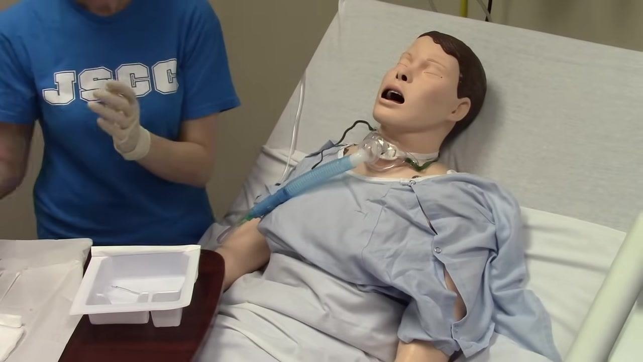 Sterile-gloved hands prepare a fenestrated trach dressing at the bedside of a mannequin patient with a tracheostomy. The sterile tray and supplies are visible on the table.