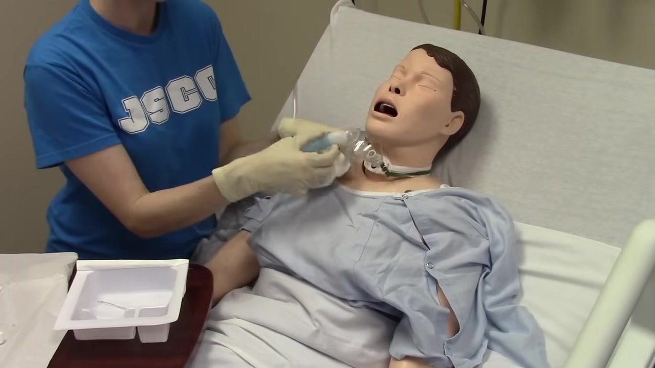 Close-up of the mannequin's neck and chest area, showing the tracheostomy tube, fenestrated dressing, and oxygen mask being adjusted and secured by gloved hands.
