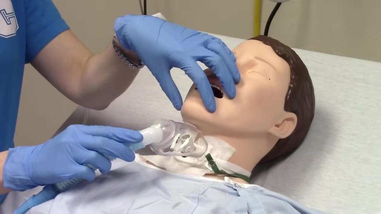 Gloved hands demonstrate the trach collar (oxygen mask) positioned over the tracheostomy site of a medical mannequin lying in a hospital bed, mouth open, with visible trach tube and collar.