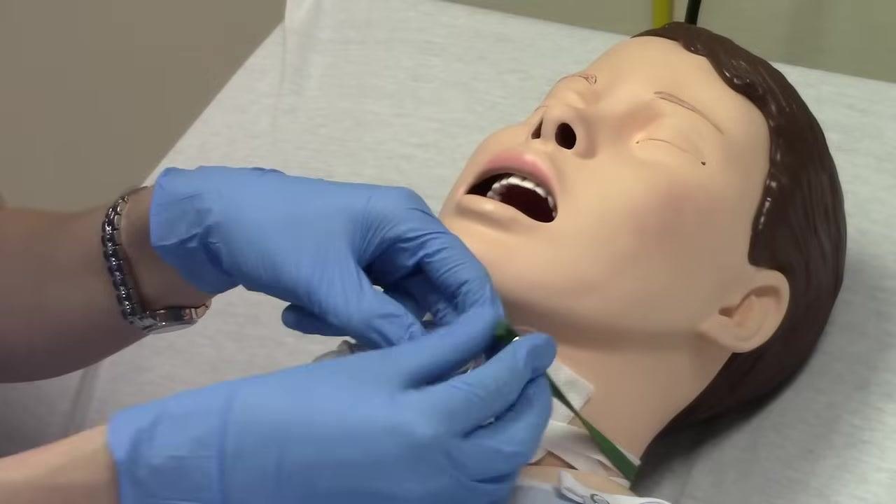 Gloved hands pointing to the size marking on the trach tube, confirming it is a number six. The trach tube and ties are clearly visible on the mannequin.