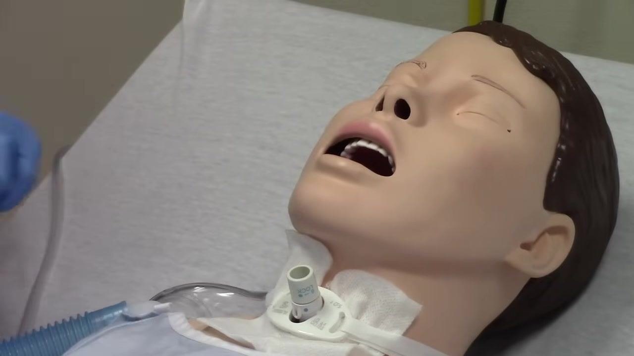 Mannequin with trach tube exposed, inner cannula removed, and gloved hand holding the oxygen tubing nearby.