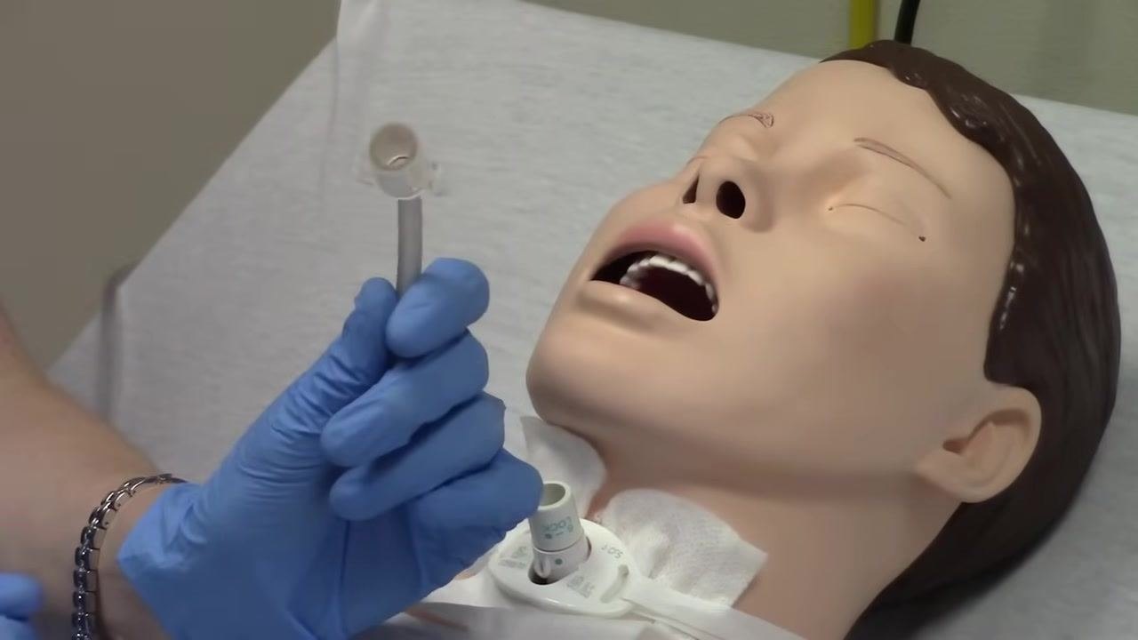 Gloved hand holding a disposable inner cannula with visible side flanges next to the tracheostomy site of a medical mannequin lying in a hospital bed with mouth open and trach tube exposed.