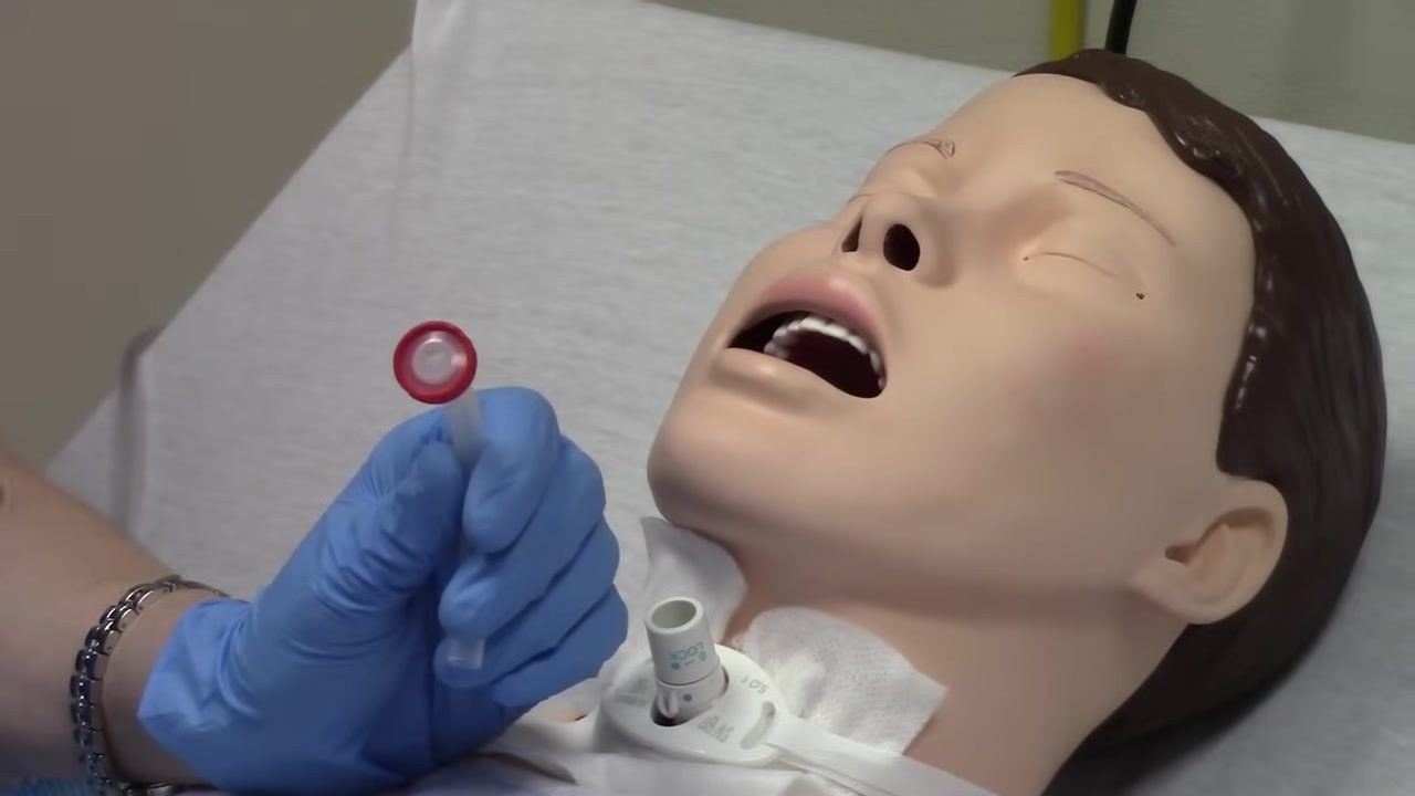 Gloved hand holding a red-capped trach button next to the mannequin's trach site, demonstrating the device used to block the trach.