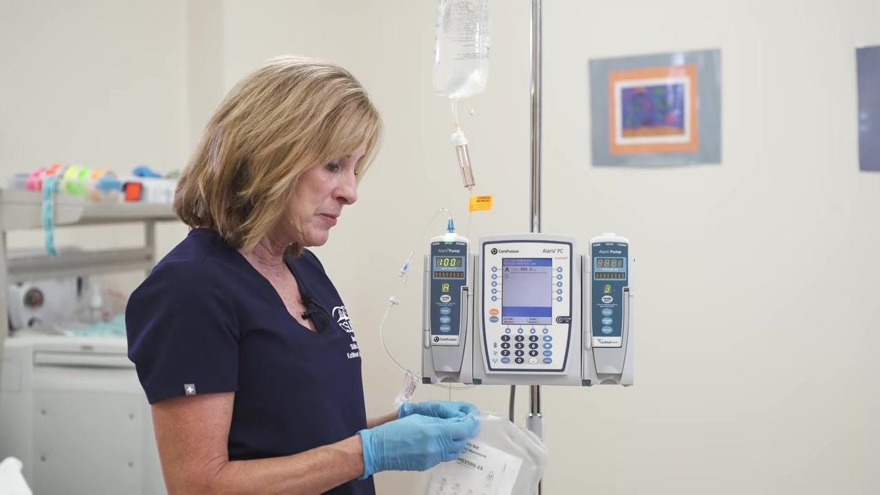 A gloved hand holds the secondary IV tubing package next to the Alaris setup.