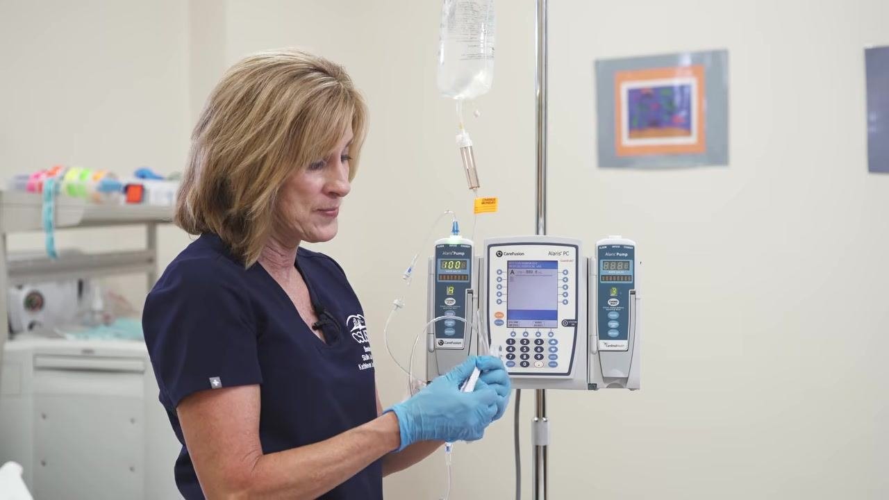 A gloved hand prepares to hang and prime the secondary tubing above the primary infusion setup.