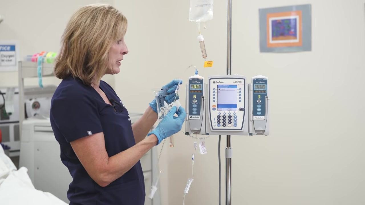A gloved hand holds the end of the secondary IV tubing near the primary setup, preparing to connect it to the correct port.