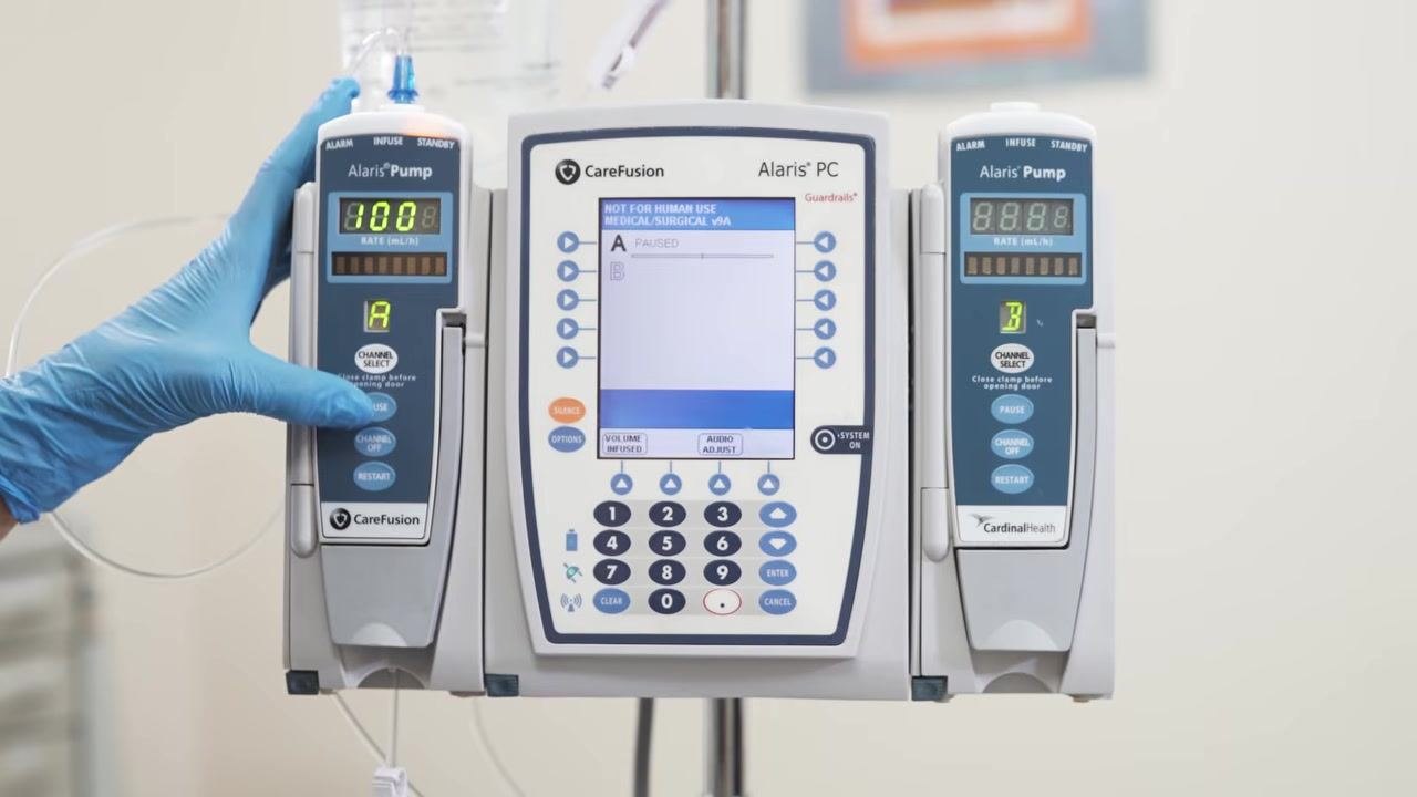 A gloved hand presses the "Channel Select" button while the pump display reads "PAUSED".