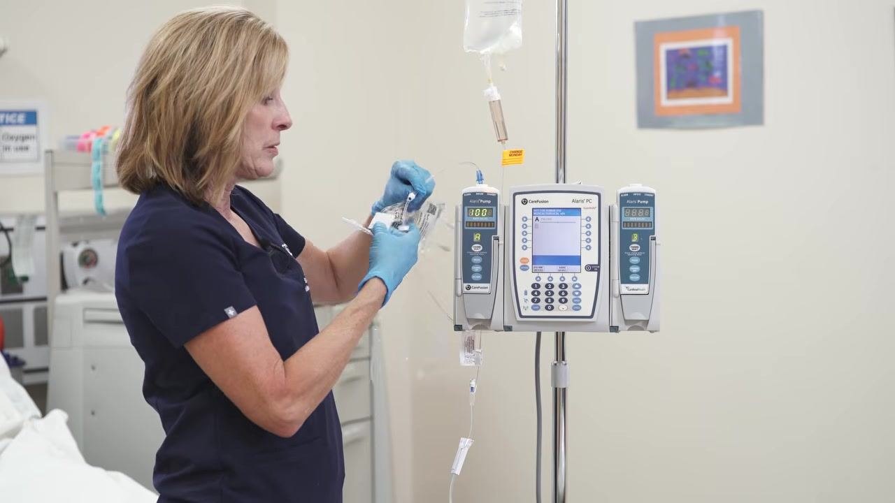 A gloved hand holds an alcohol swab, preparing to cleanse the secondary port above the pump.
