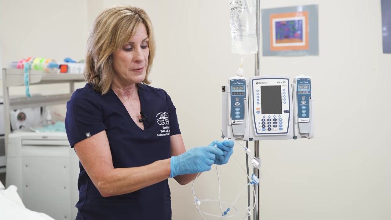 A gloved hand holds the IV bag and the sterile tubing spike, preparing to insert it into the bag.