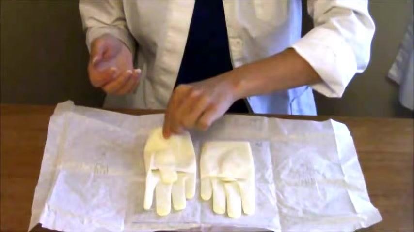 The non-dominant hand grasps the inside cuff of the first glove with the thumb tucked in, demonstrating contamination-free technique.