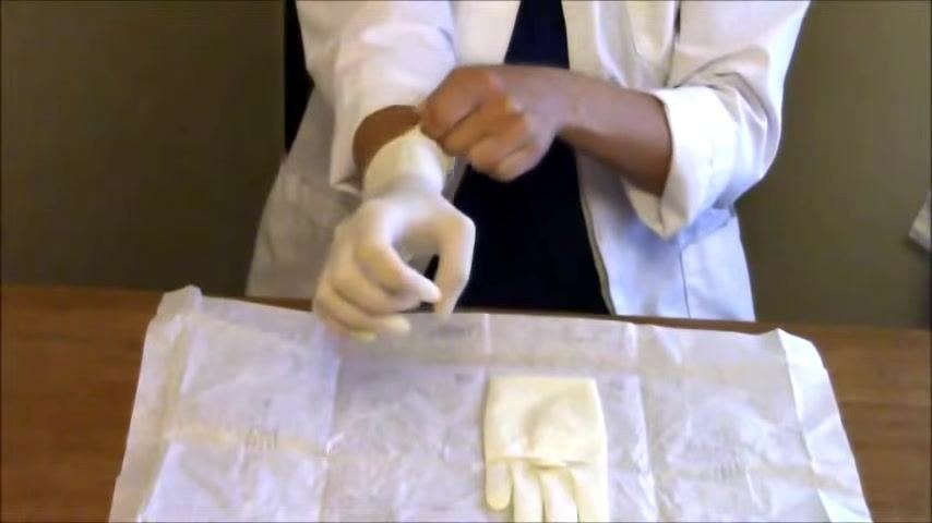 The dominant hand is pulled into the first glove using the inside cuff, with the thumb tucked in to preserve sterility.