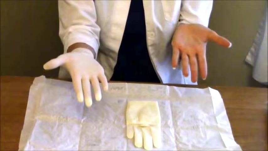 A sterile gloved hand slides its fingers underneath the cuff of the second glove, preparing to glove the non-dominant hand.